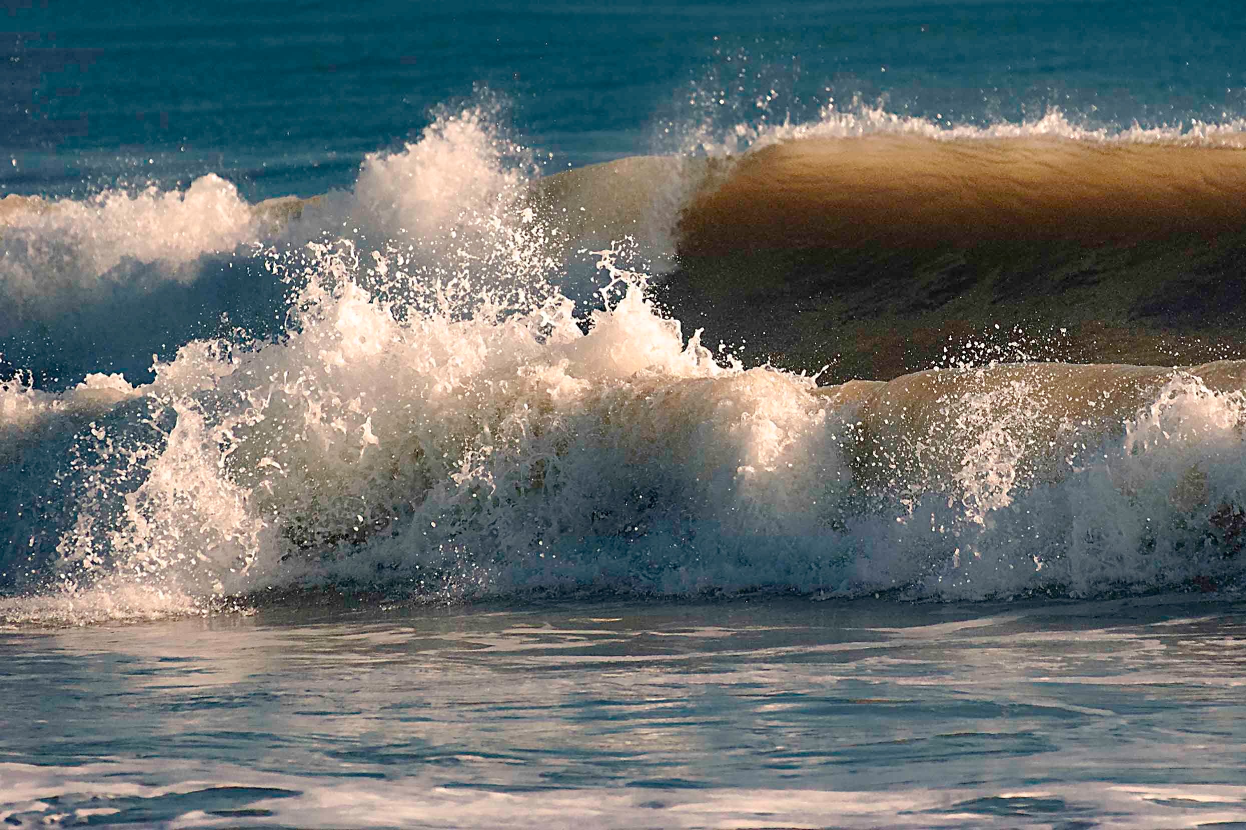 A wave cresting and breaking in bright afternoon light with white spray against deep blue water