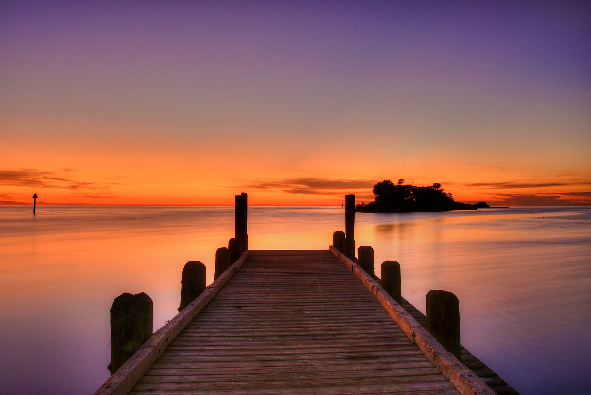 A wooden dock stretching into glassy Gulf waters at twilight, the sky fading from deep orange at the horizon through lavender and purple overhead