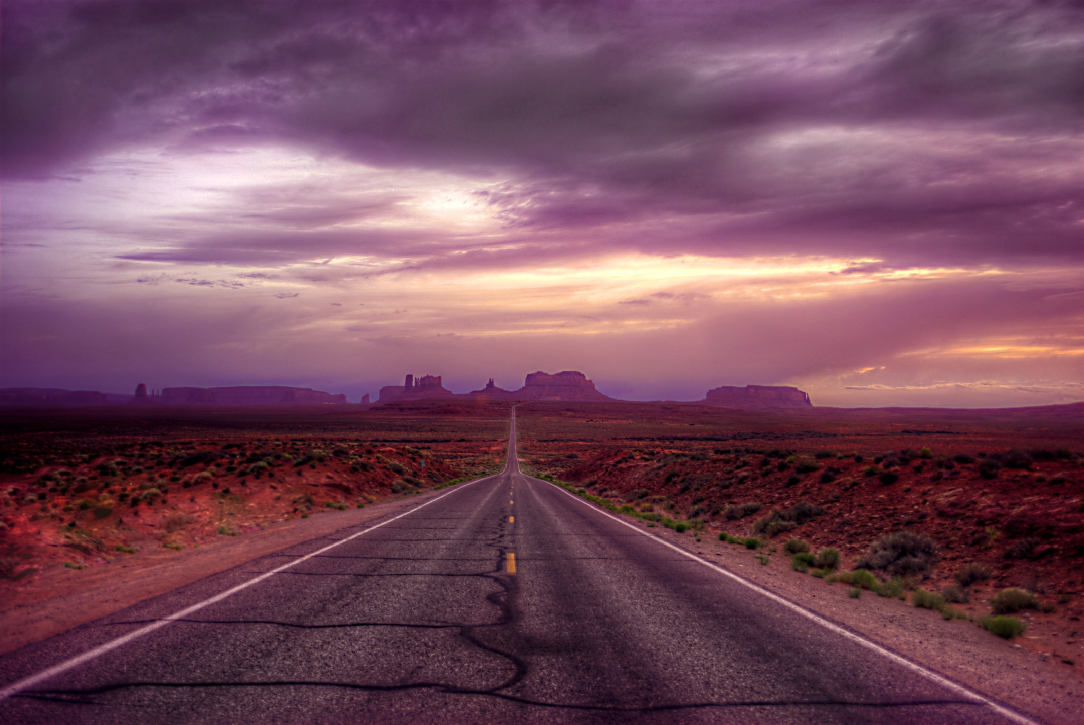 A desert highway stretching toward the sandstone buttes of Monument Valley beneath a dramatic purple and gold sunset sky