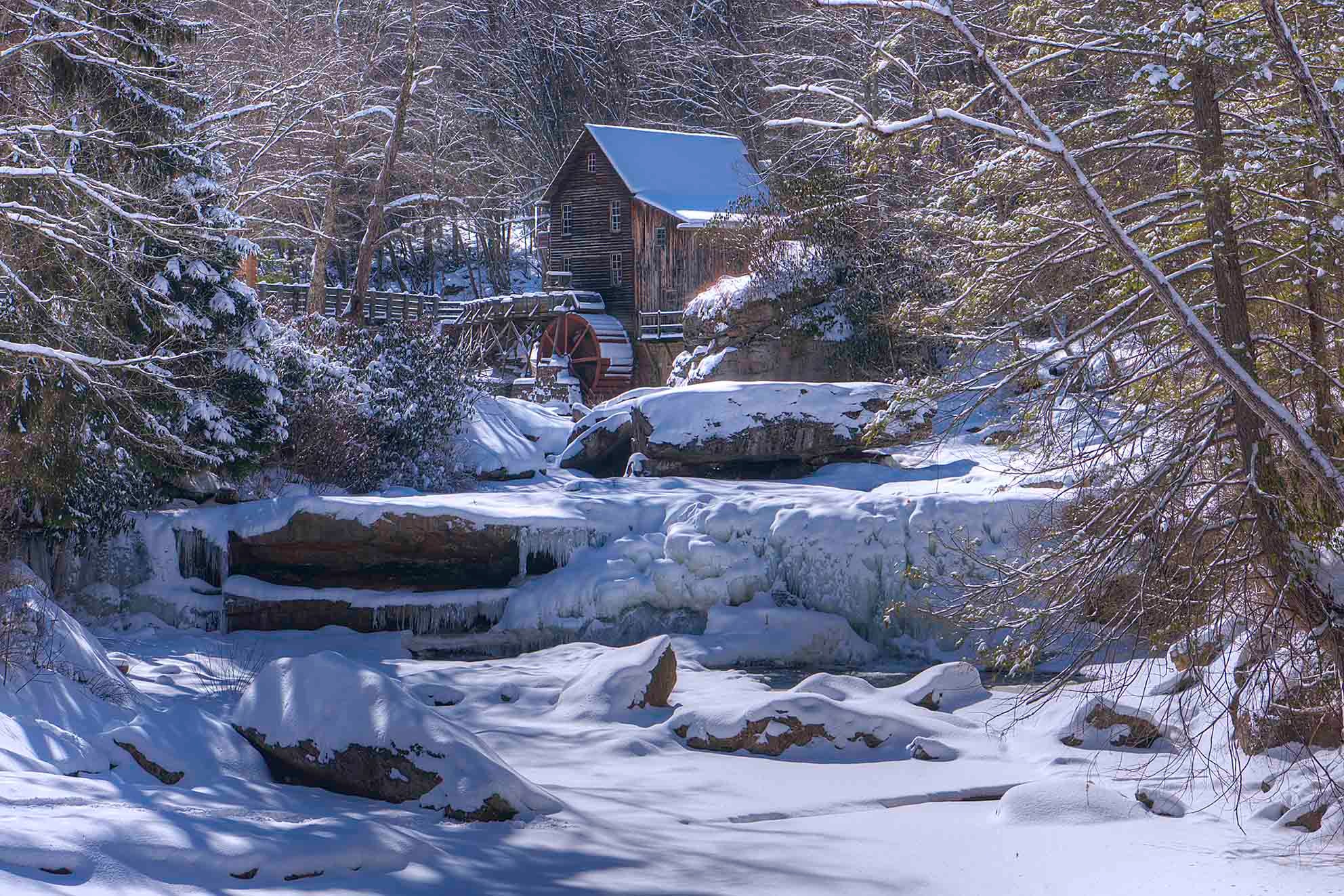 Snow-covered grist mill with red water wheel beside a frozen stream in a winter forest