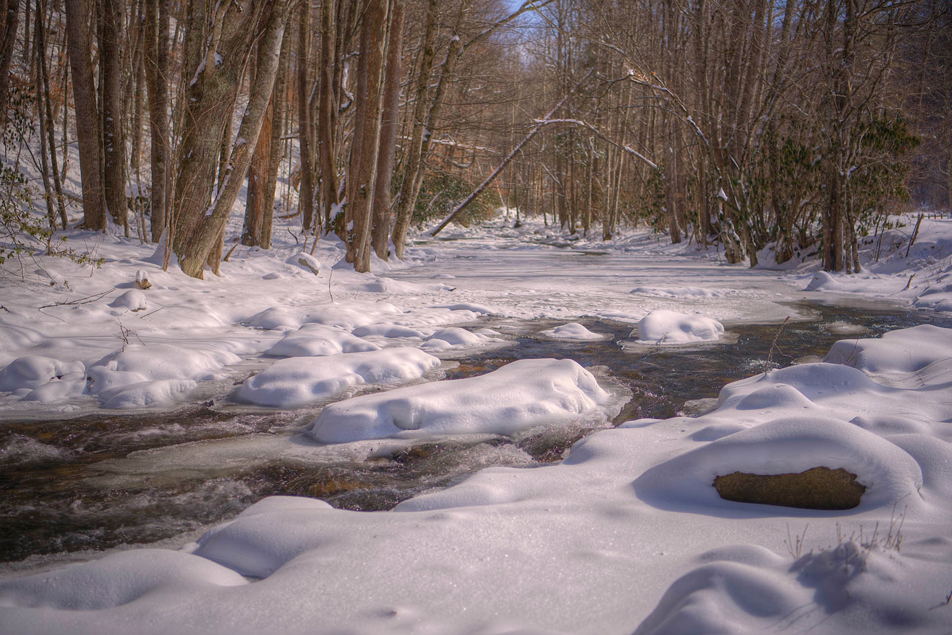 Snow-capped boulders lining a gently flowing stream through a bare winter forest