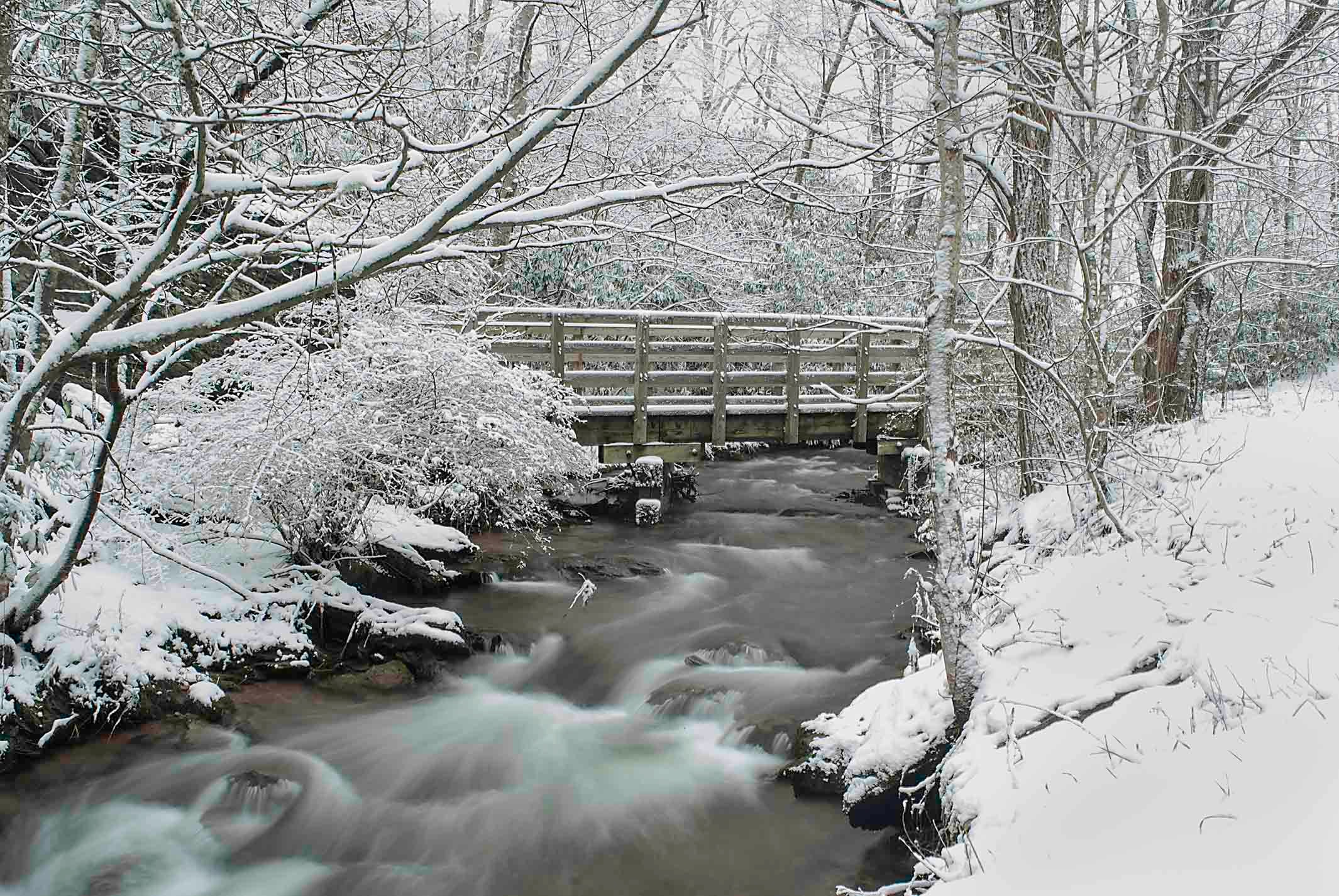 Snow-covered wooden bridge over a rushing stream in a bare winter forest in the Virginia Highlands