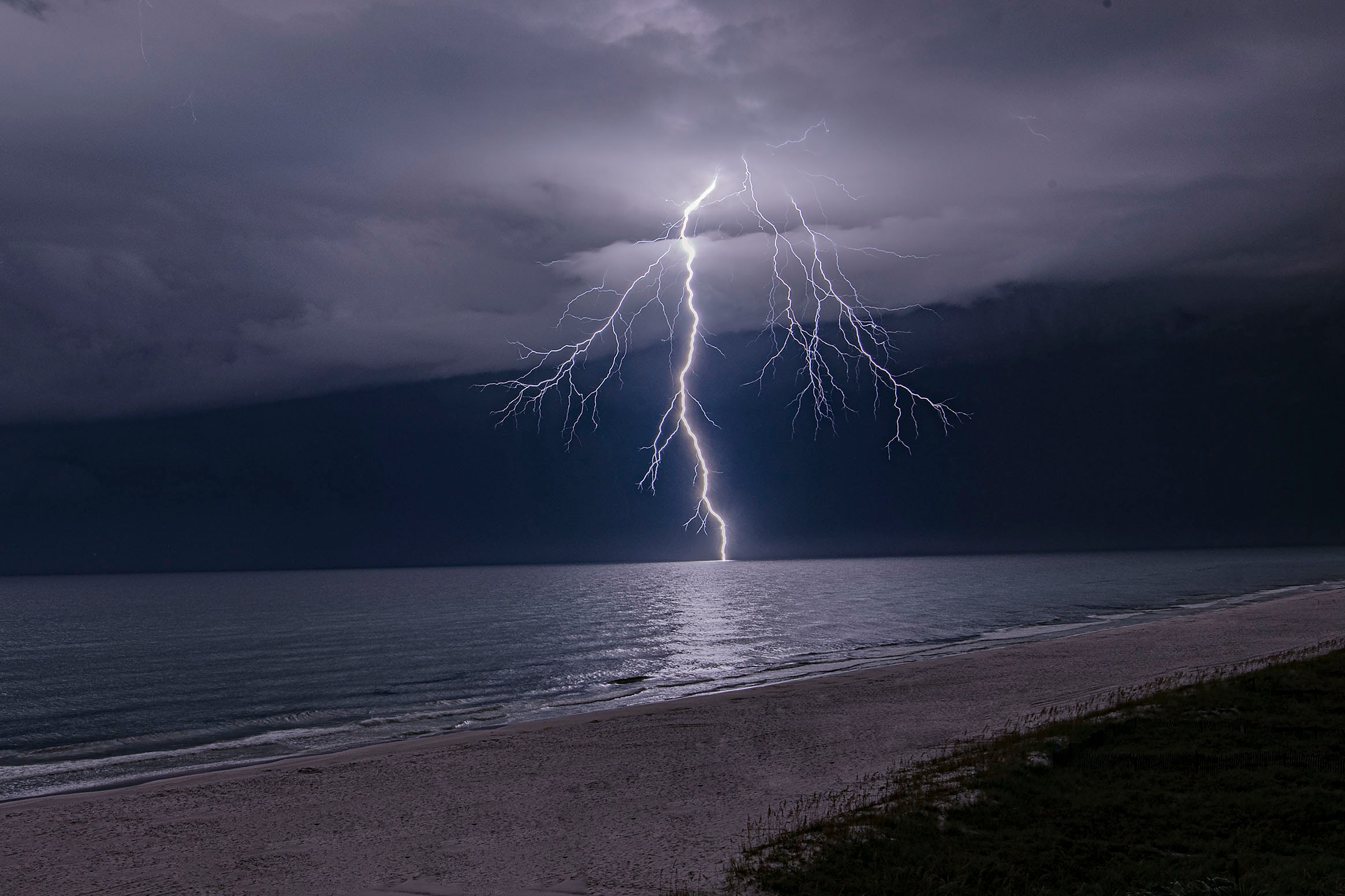 A single massive lightning bolt striking the ocean from a dark storm cell, its light reflected in a long column on the water