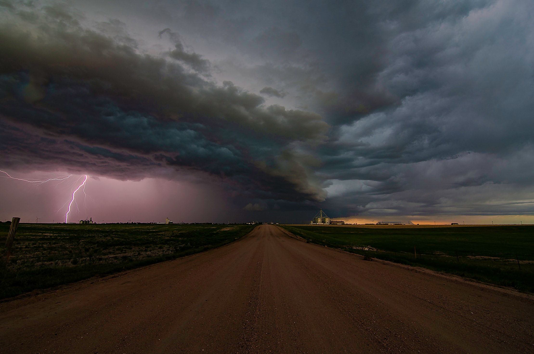 A supercell thunderstorm with a shelf cloud and lightning bearing down on a flat prairie road toward grain silos at dusk