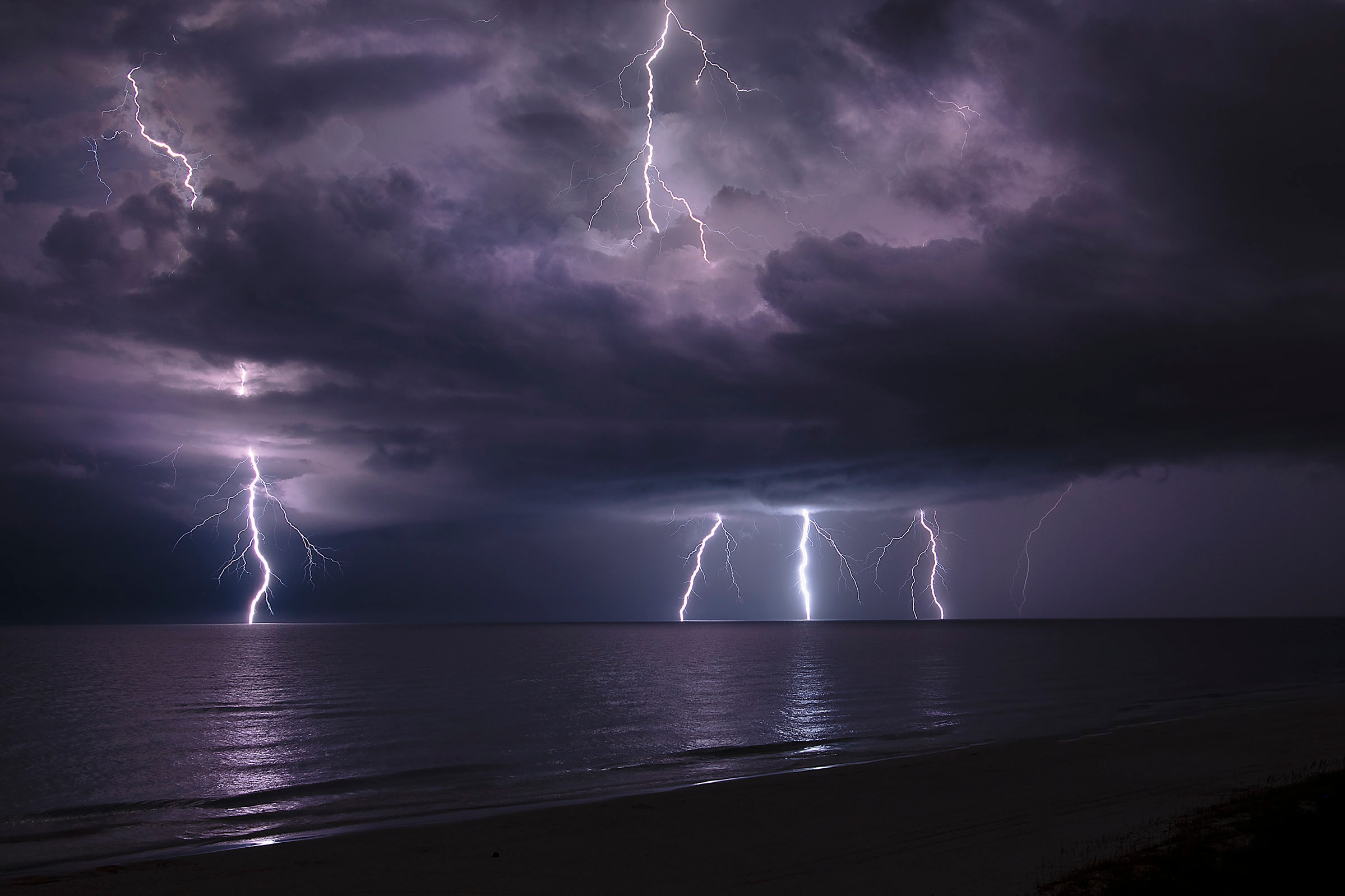 Lightning bolts striking the ocean across a wide panoramic view from a dark beach at night
