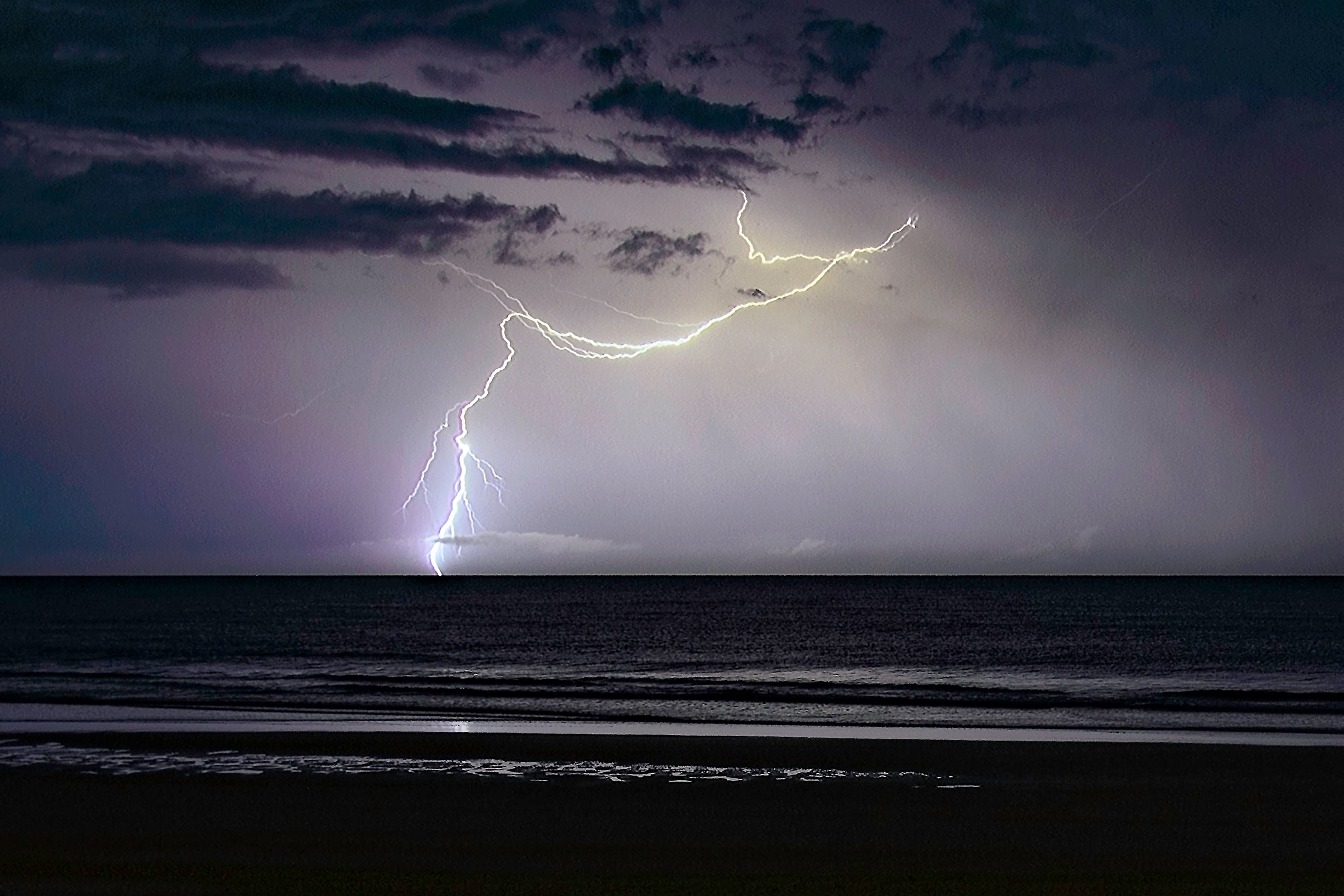 A dramatic branching lightning bolt over the dark ocean at night, illuminating the beach and water in purple and white light