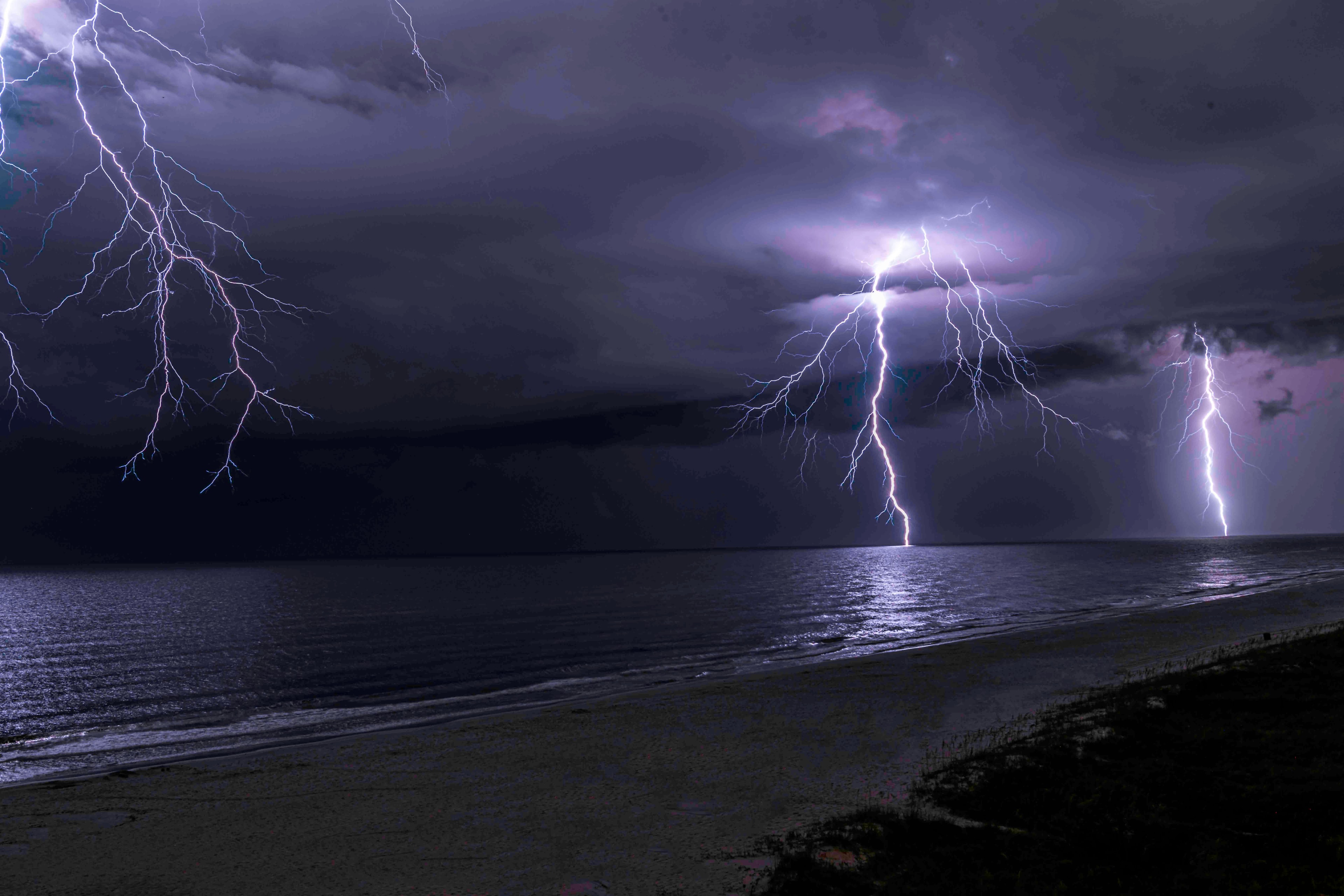 Lightning bolts striking the ocean from a dark storm cell, reflected in the wet sand of a Florida beach